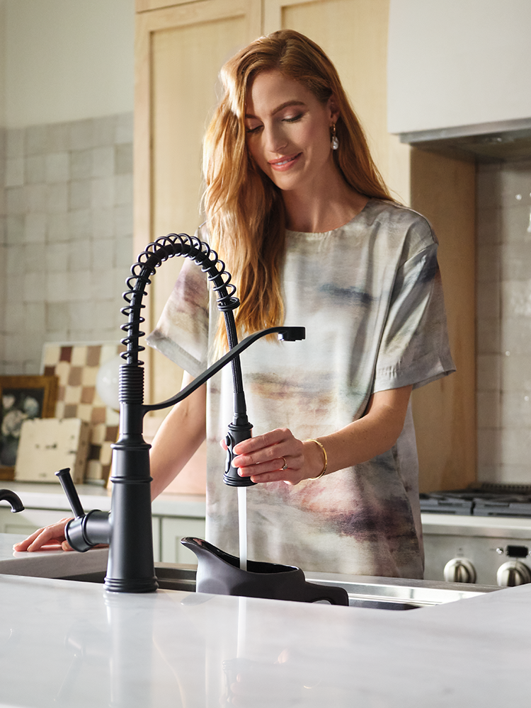 A woman with long red hair stands at a kitchen sink, holding the trigger sprayer of a matte black faucet. She is wearing a short-sleeve t-shirt with a gray and white abstract print.