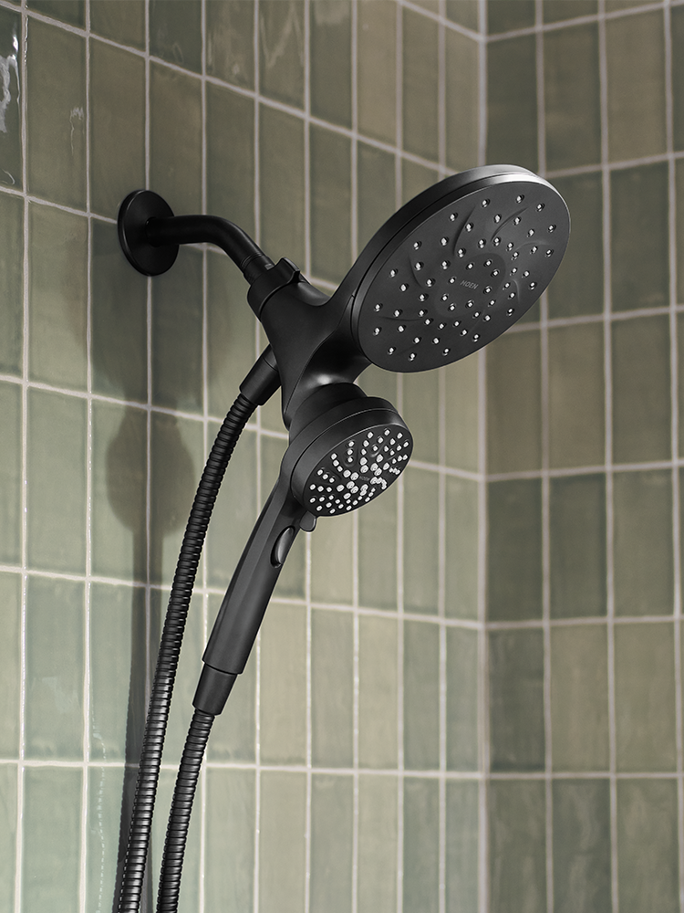 A matte black dual-head shower system with a wall-mounted round shower head, flexible hose, and handheld shower attachment, shown installed against a green tiled bathroom wall.