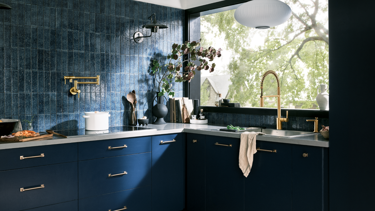 A modern kitchen interior with dark blue cabinets, a matching blue tiled backsplash, a large window with outdoor greenery visible, and gold-toned fixtures including a faucet and drawer pulls.