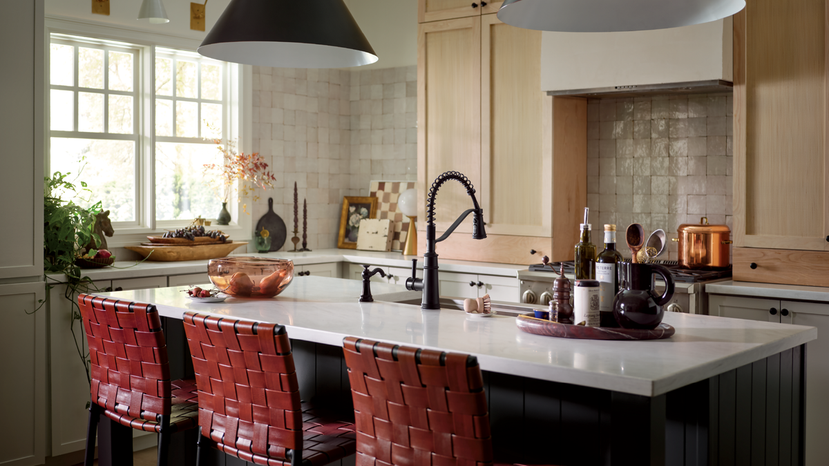 A modern kitchen island with a white countertop, black gooseneck faucet, and three red woven bar stools, set in a bright kitchen with light wood cabinets and a tiled backsplash.