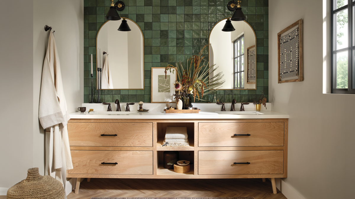 A double-sink bathroom vanity in a real bathroom setting, with a light natural wood cabinet, white countertops, two arched mirrors, green tiled backsplash, hanging towels, and decorative plants.