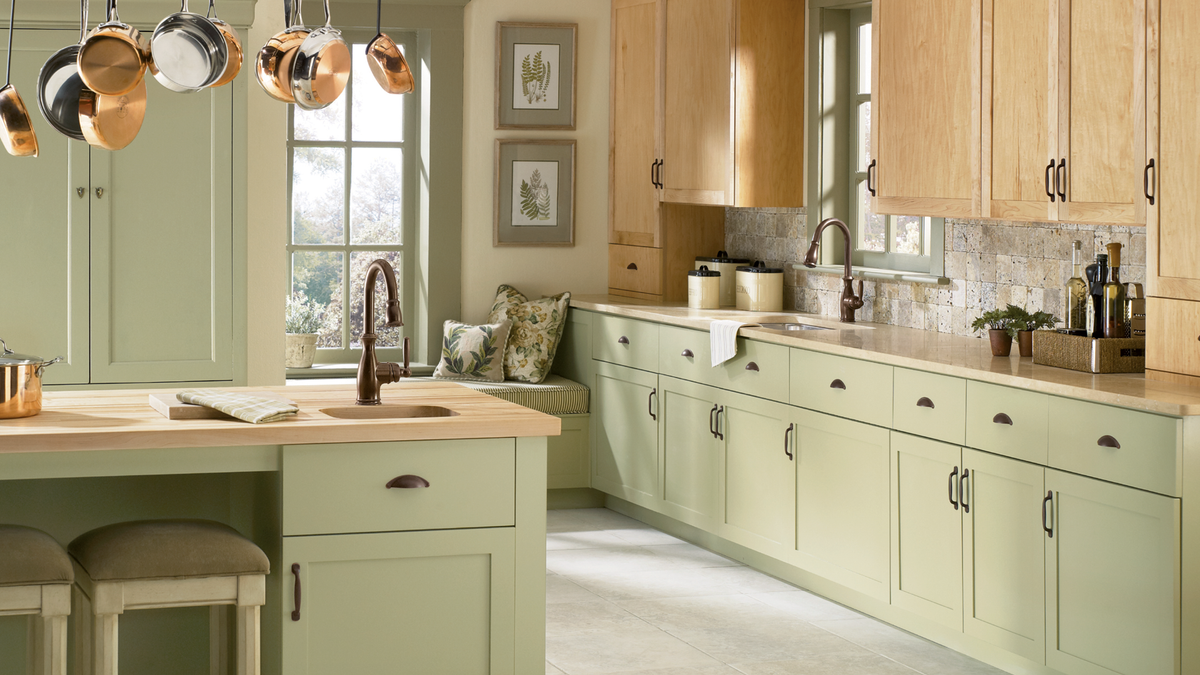 A bright, modern kitchen featuring sage green cabinets and island, light wood upper cabinets, beige countertops, and copper pendant lights hanging over the island.