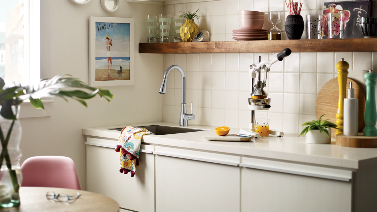 A bright, modern kitchen interior with cream-colored cabinets, a tiled backsplash, a chrome faucet, and open wooden shelves holding dishes and glassware. A framed beach print hangs on the wall, and a pink chair is partially visible in the foreground.
