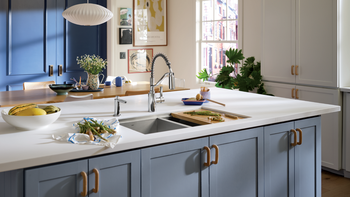 A modern kitchen island with light blue cabinets, a white countertop, a double-bowl sink, and brass handles, shown in a bright, styled kitchen interior.
