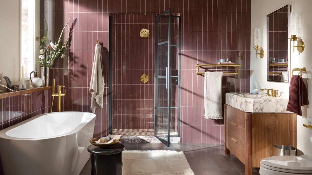 A luxury bathroom interior featuring a freestanding white bathtub, a walk-in shower with a glass door, a wooden vanity with a marble countertop, and rich burgundy wall tiles. Gold fixtures and brass lighting fixtures are prominently visible throughout the space.