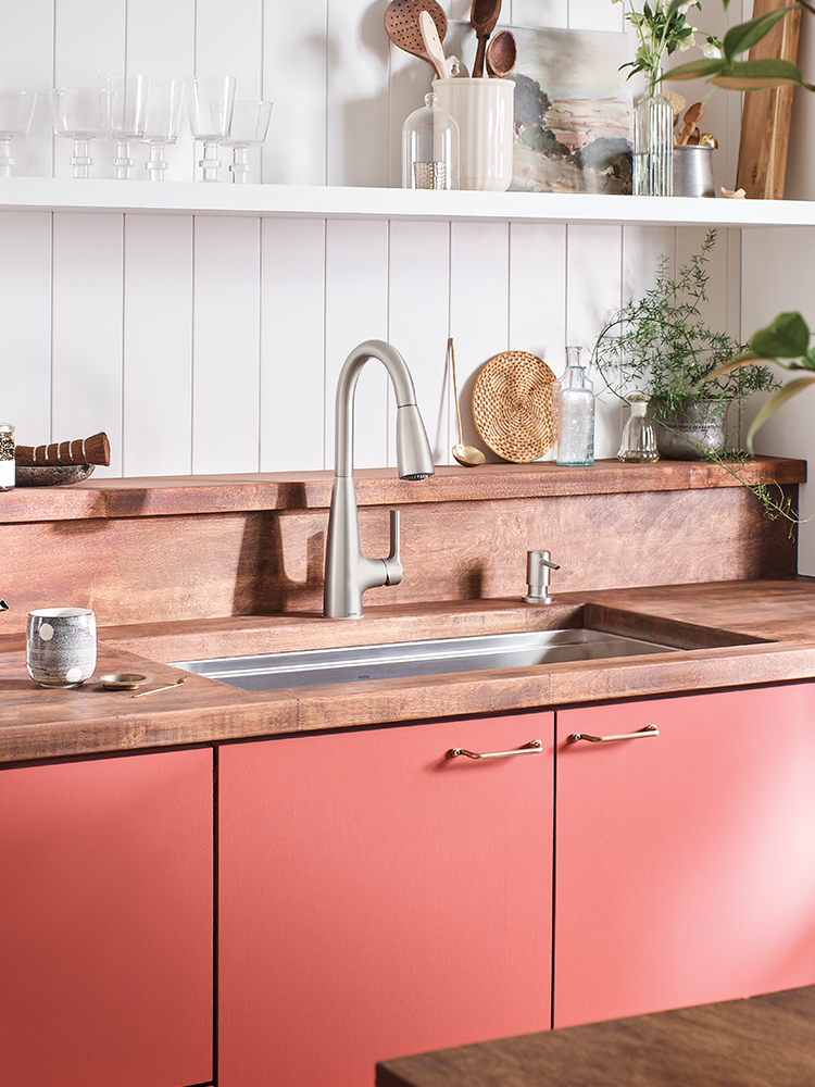 A modern kitchen scene featuring a brushed nickel single-handle faucet with a pull-down sprayer, installed in a stainless steel sink set into a warm wood countertop and backsplash. The lower cabinets are a coral pink color, and the background consists of white shiplap walls with open shelves holding glassware and decorative items.