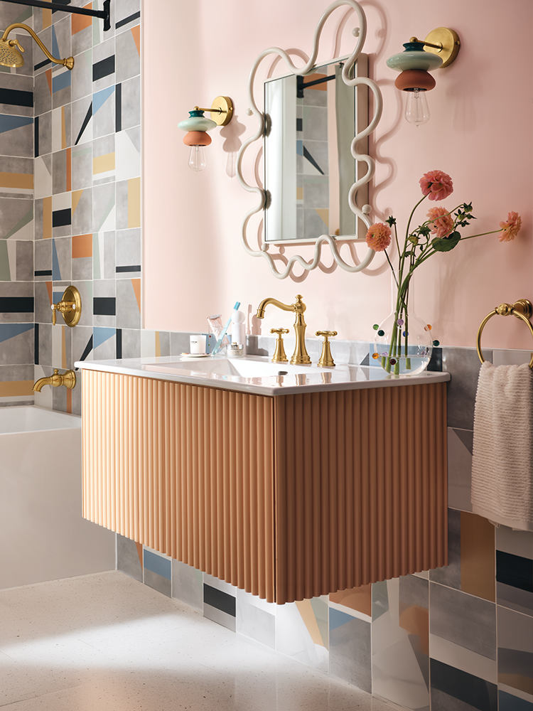 A modern bathroom vanity with a fluted peach-orange cabinet, white countertop, and gold faucet, set against a pink wall with an ornate mirror and colorful geometric wall tiles.