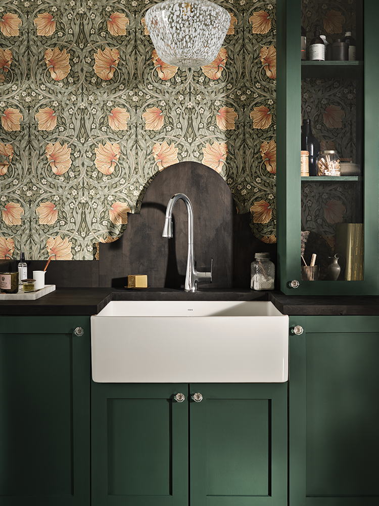 A white farmhouse-style undermount sink set into dark green kitchen cabinets, with a curved dark backsplash, a chrome gooseneck faucet, and ornate floral wallpaper behind it.