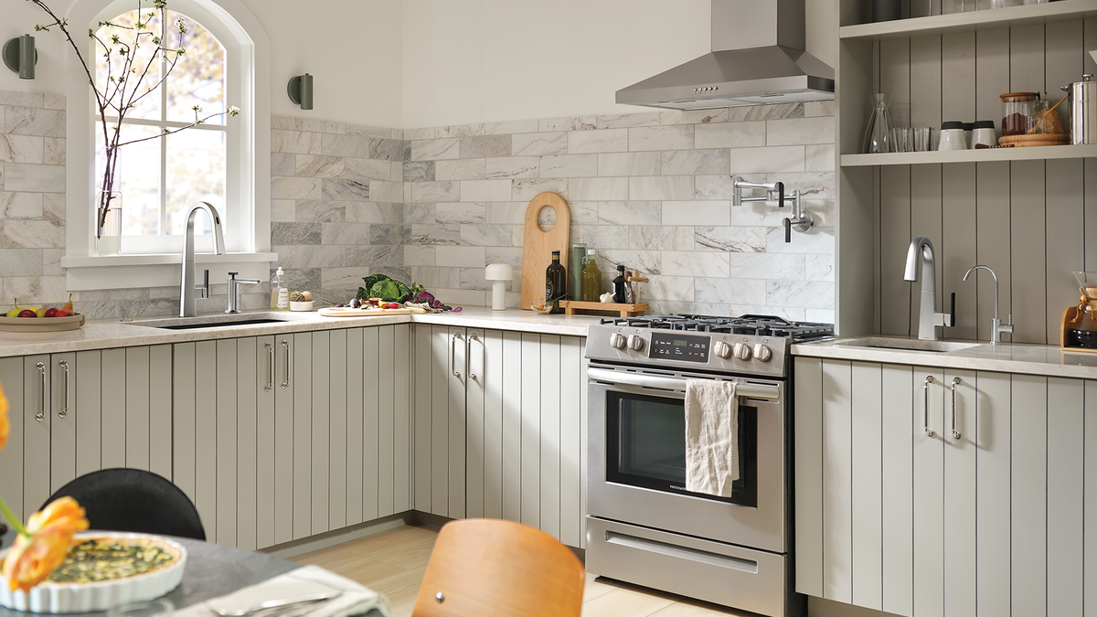 A modern L-shaped kitchen featuring light gray shiplap cabinets, a gray marble backsplash, stainless steel appliances including a gas stove and range hood, and open shelving on the upper right.