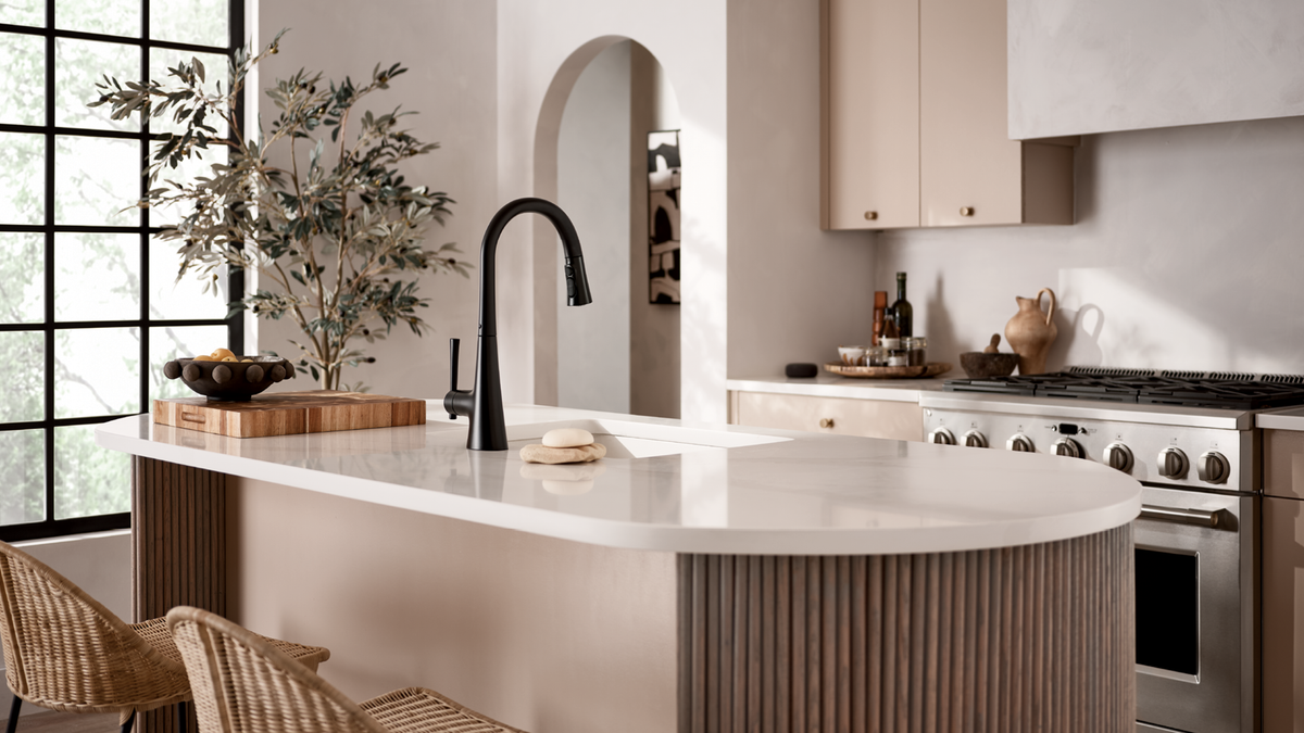 A modern kitchen island with a white curved countertop, fluted wood base, black gooseneck faucet, and woven bar stools in a bright neutral kitchen.