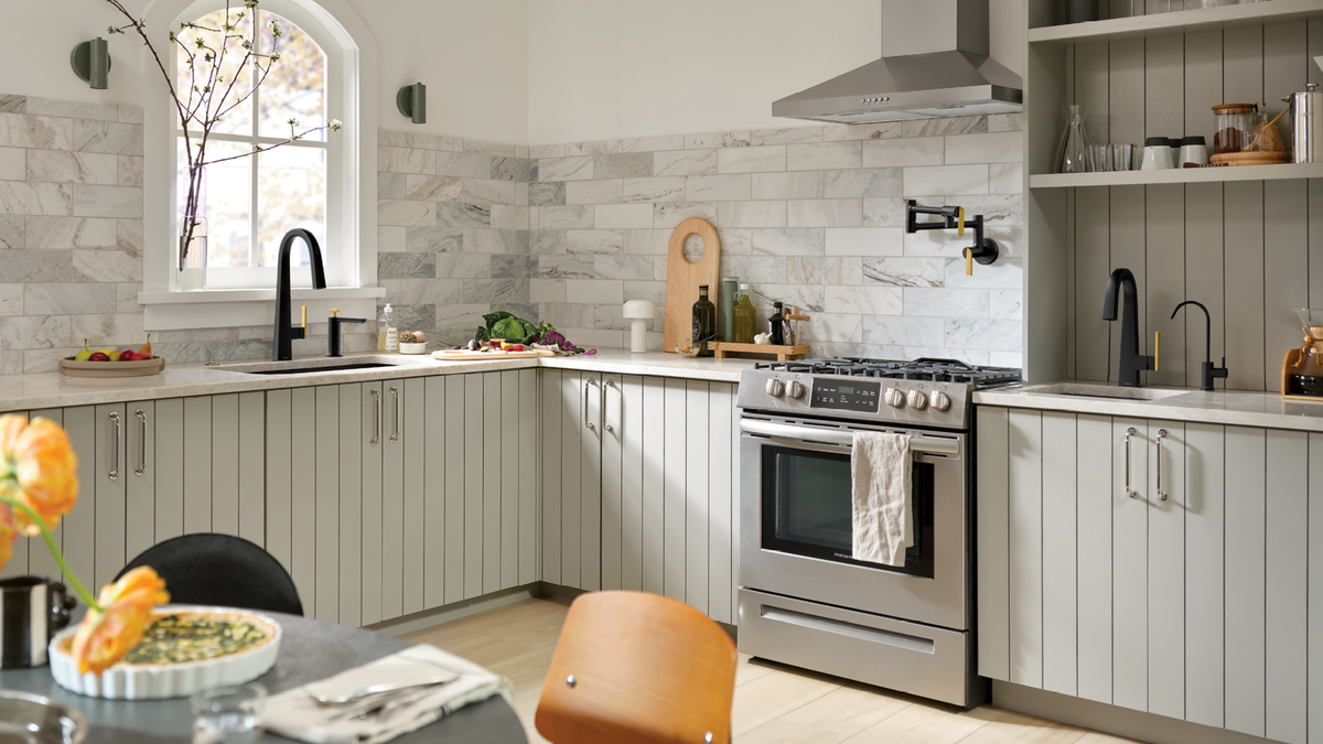 A modern kitchen interior with light gray shiplap cabinets, white marble tile backsplash, stainless steel gas range, black faucets, open shelving, and a window with bare branches.