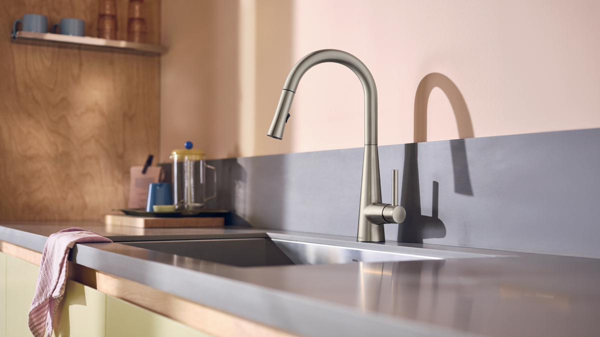 A modern matte silver pull-down kitchen faucet installed over a double-basin stainless steel sink on a gray countertop, with a warm peach-pink wall and wooden backsplash in the background.