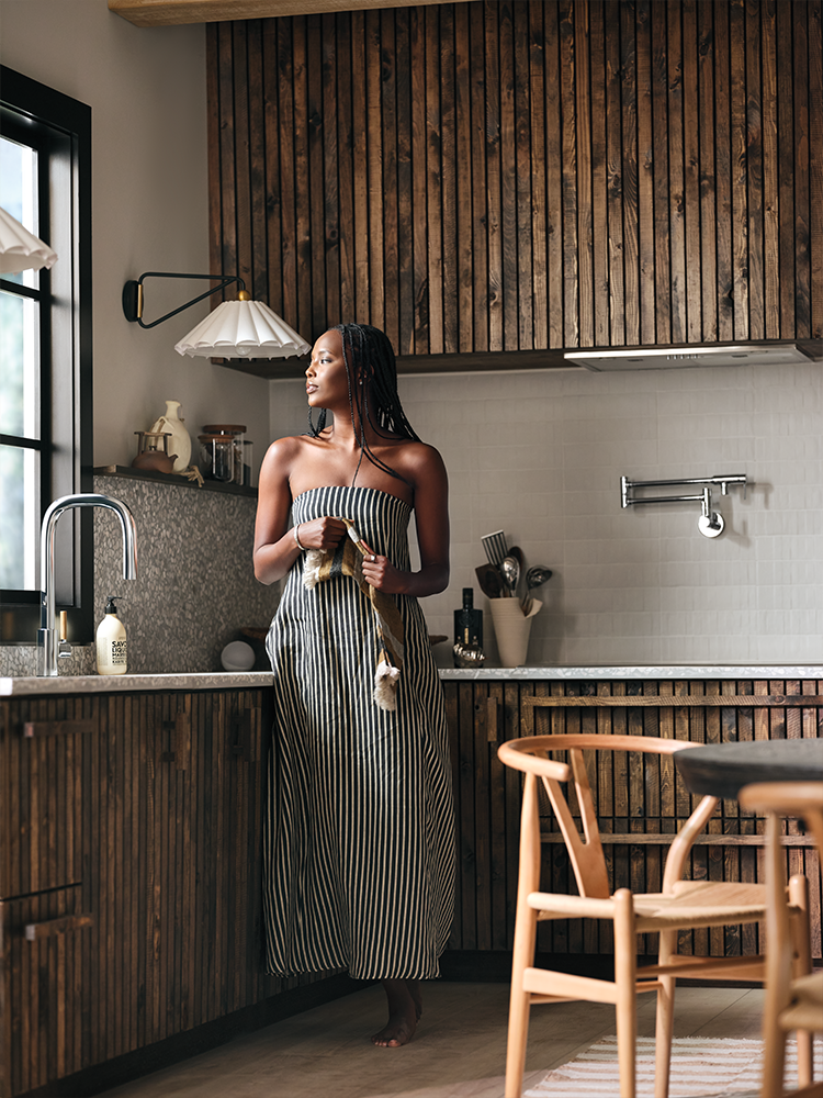 A woman wearing a black and white vertically striped strapless maxi dress stands in a rustic kitchen with wood-paneled walls and cabinets, holding a small woven bag.