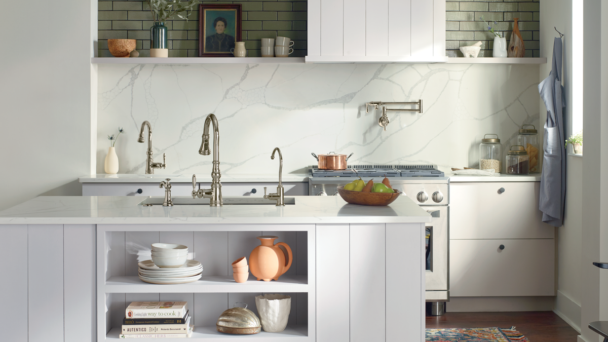 A modern kitchen interior with white cabinetry, a white marble backsplash, open shelving with dishes and decorative items, a gas stove, and a hanging apron near the window.