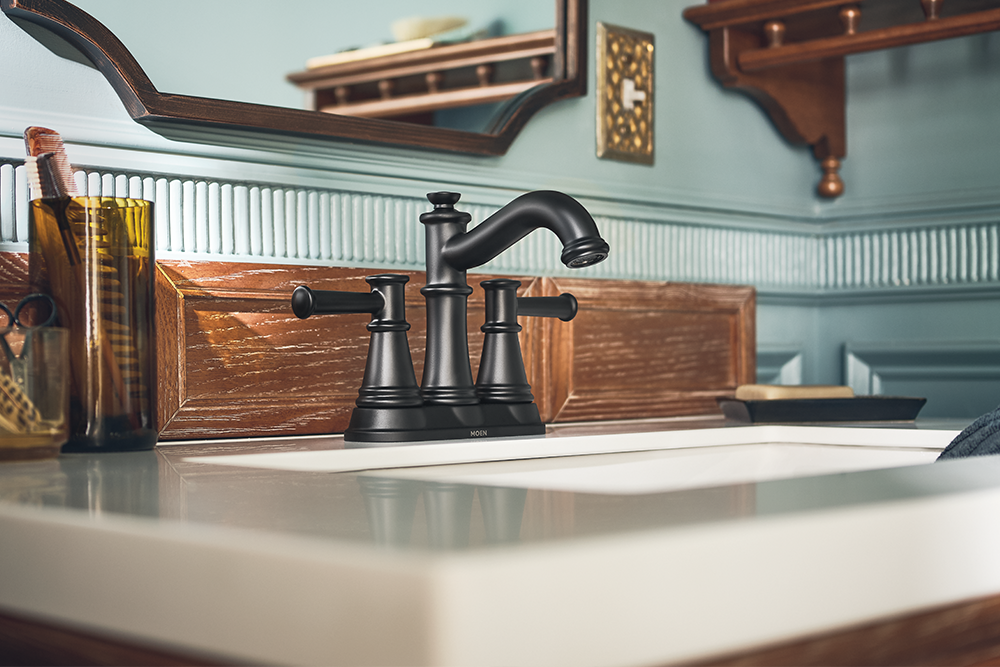 A matte black three-hole bathroom faucet set installed on a white sink countertop, with a wooden backsplash, light blue paneled wall, and mirror in the background.