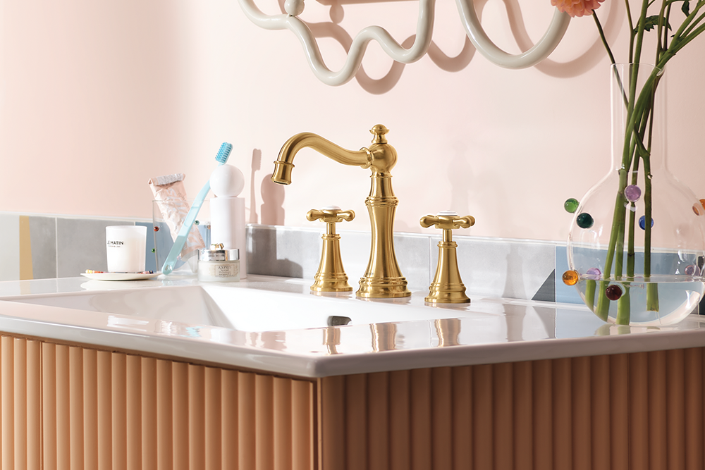 A polished gold three-hole bathroom faucet set installed in a white sink, set against a fluted light brown vanity and a soft pink wall.