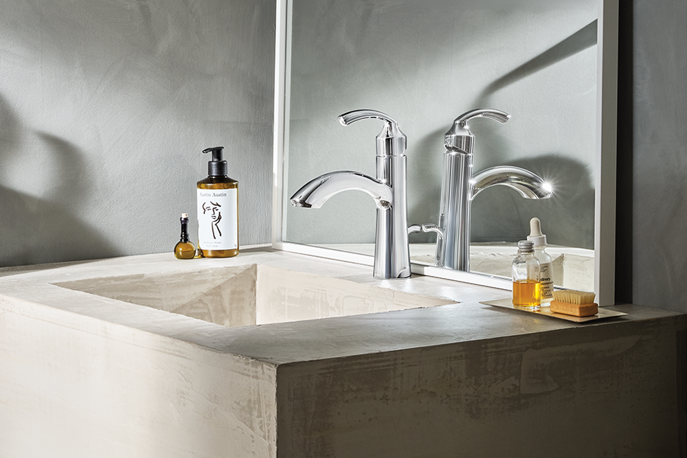 A modern chrome bathroom faucet installed in a concrete sink, shown with a matching mirror and bathroom accessories in a minimalist interior.