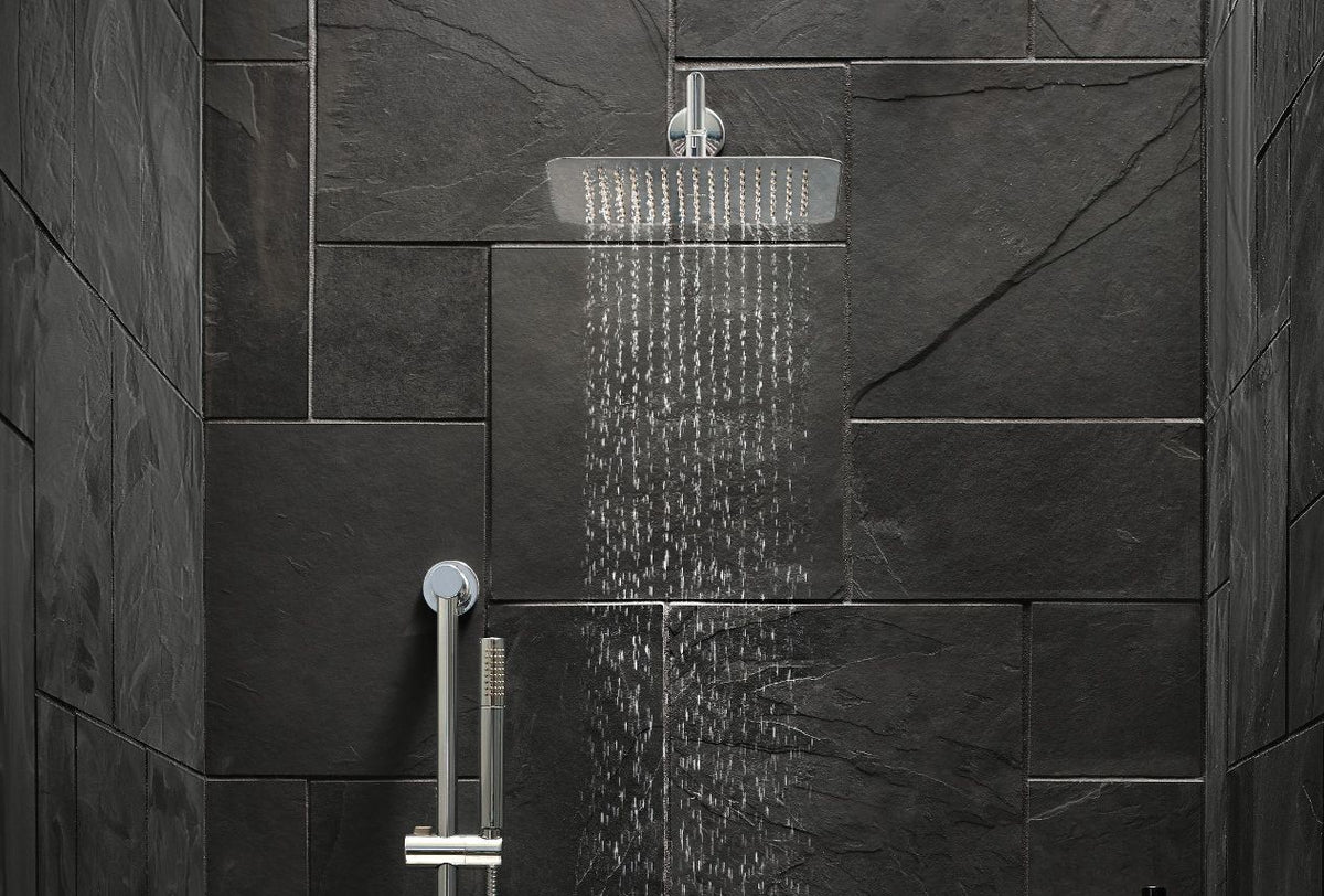 A modern square rain shower head with water running from it, mounted on a wall with dark charcoal stone tiles. A chrome handheld shower attachment is visible below.