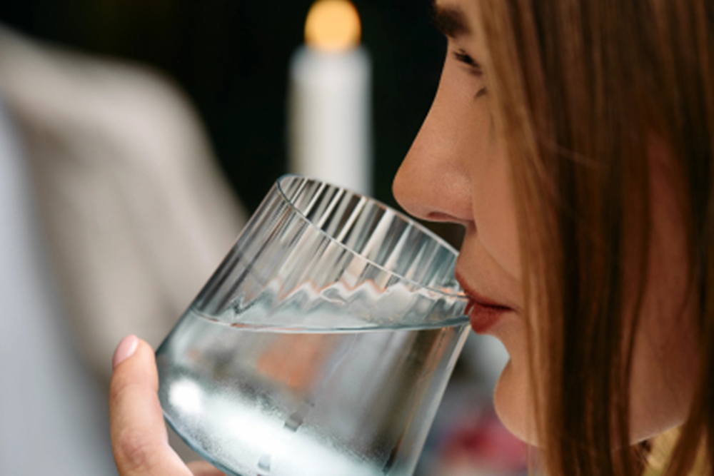 A close-up lifestyle shot of a woman drinking from a clear ribbed glass tumbler, with a softly blurred candle in the background.